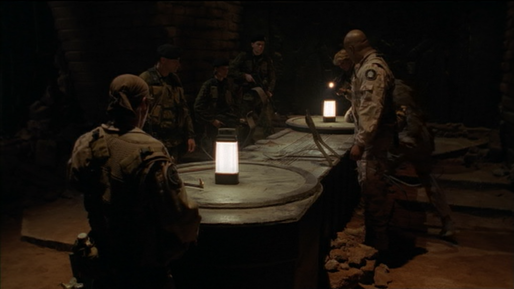 SG1 und das russische Team stehen um einen Goa'uld-Sarkophag herum. Auf dem Sarkophag stehen Lampen, der Rest des Raumes ist sehr dunkel.